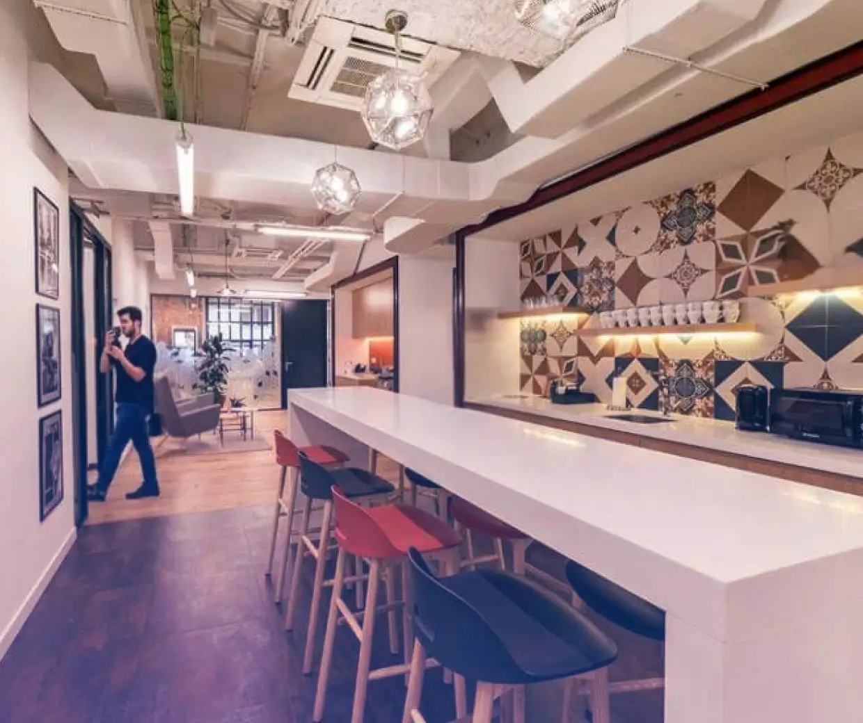 Modern office kitchen area with bar stools, geometric tile backsplash, and a man walking with coffee